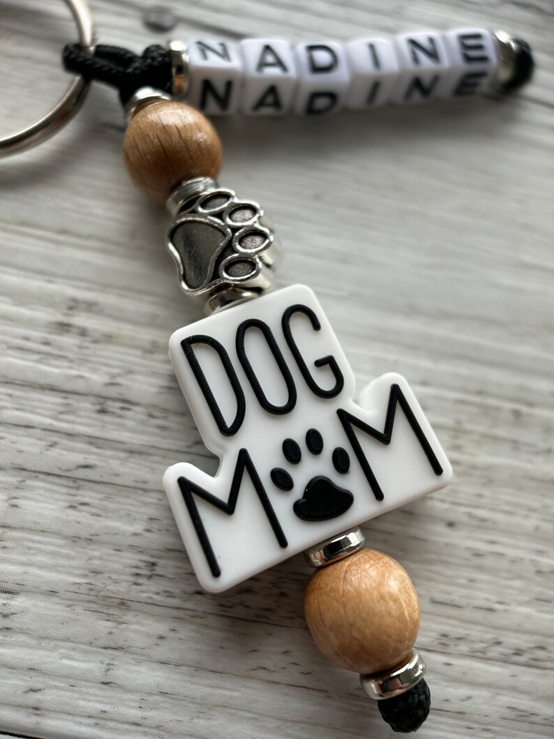 May include: White keychain with black text that reads "Dog Mom" and a black paw print. The keychain has a silver paw print charm and wooden beads.