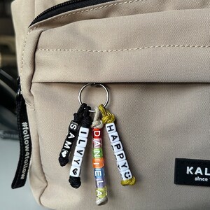 May include: A keychain with personalized name beads attached to a beige backpack. The keychain features the names "SAM", "ILVY", "DANIELA", and "HAPPY" spelled out with colorful letter beads. The text "In meinem Shop findest du noch viele weitere Varianten der Namensanh&auml;nger :)" is visible.
