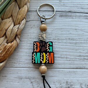 May include: A black and colourful keychain with the words "Dog Mom" and a paw print. The keychain has wooden beads and a silver key ring.