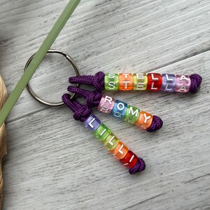 May include: Three personalized keychains with names spelled out in colorful, translucent letter beads. Each keychain has a purple knotted cord and a silver key ring. The names are "STELLA", "ROMY", and "LILLI".
