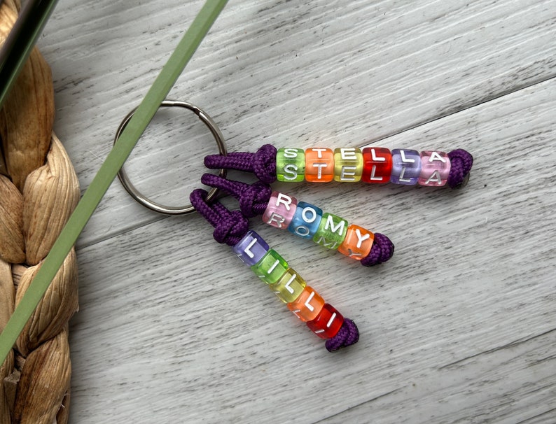 May include: Three personalized keychains with names spelled out in colorful, translucent letter beads. Each keychain has a purple knotted cord and a silver key ring. The names are "STELLA", "ROMY", and "LILLI".