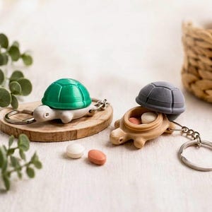 Könnte beinhalten: Zwei schildkrötenförmige Schlüsselanhänger mit Metallringen. Einer hat einen grünen Panzer und einen hellen Körper, der andere einen grauen Panzer und einen beigen Körper. Die graue Schildkröte ist offen und zeigt ein kleines Fach mit zwei Pillen.