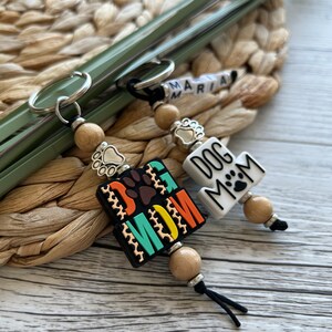 May include: Two keychains with wooden beads and metal charms. One keychain has a black and white tag that says "Dog Mom" with a paw print. The other keychain has a colourful tag that says "Dog Mom" with a paw print.