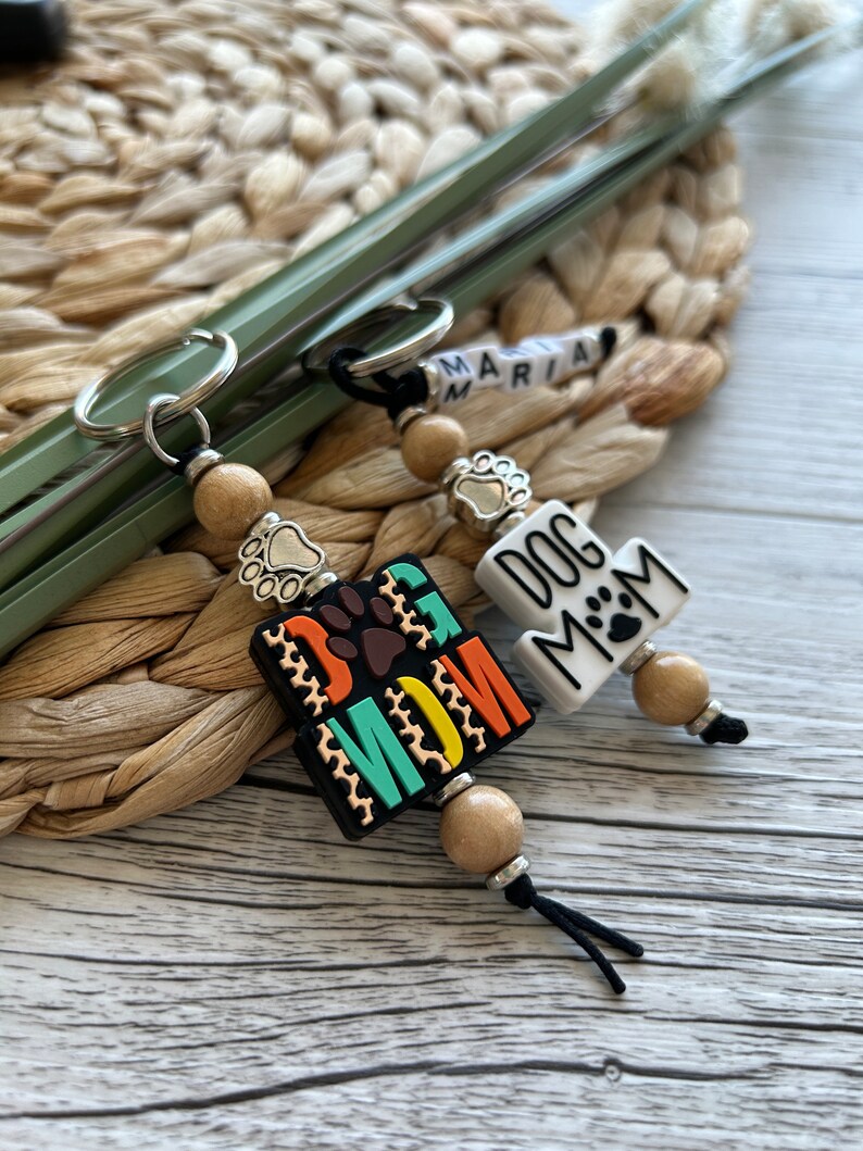 May include: Two keychains with wooden beads and metal charms. One keychain has a black and white tag that says "Dog Mom" with a paw print. The other keychain has a colourful tag that says "Dog Mom" with a paw print.