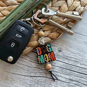 May include: A colourful keychain with the words "Dog Mom" and a paw print. The keychain is made of wooden beads and has a black tassel.