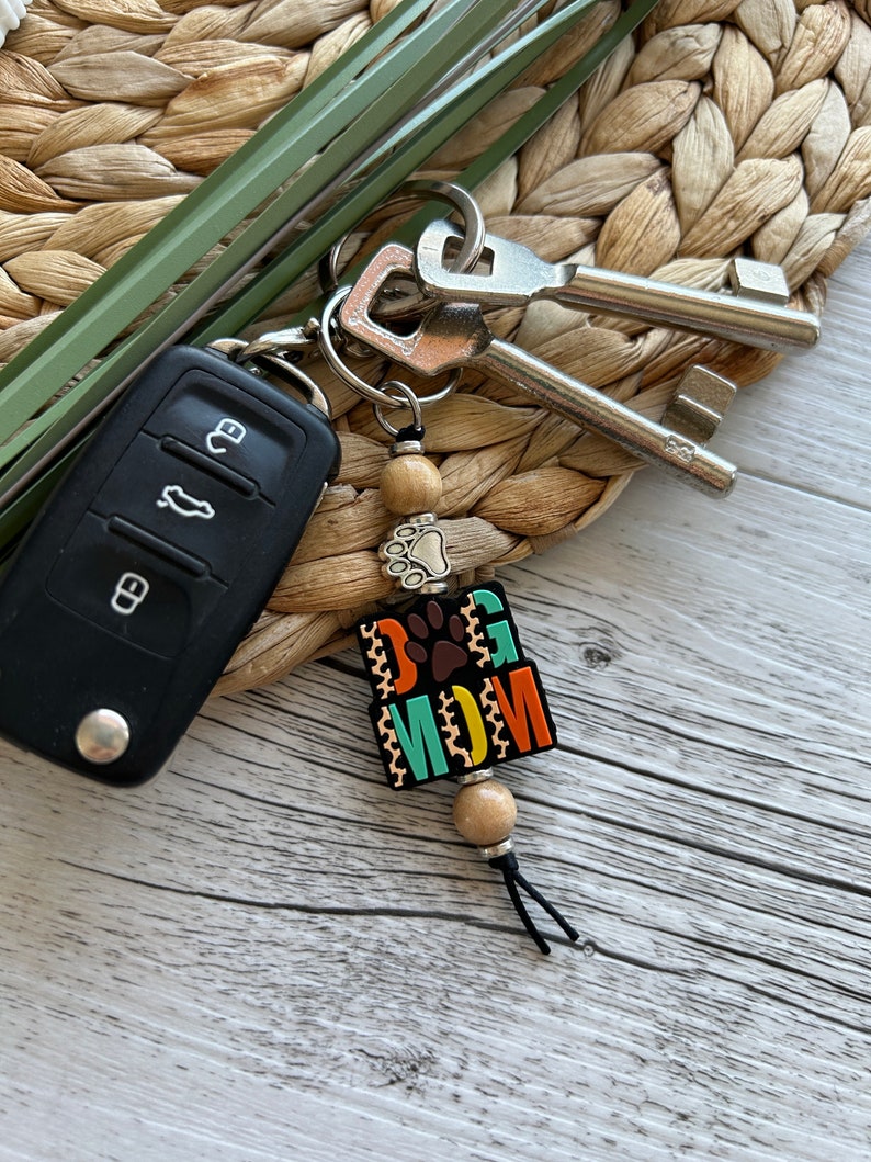May include: A colourful keychain with the words "Dog Mom" and a paw print. The keychain is made of wooden beads and has a black tassel.