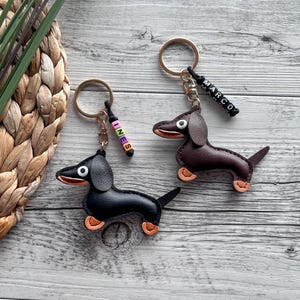 May include: Two dachshund keychains, one black and one brown, with orange accents on their paws and mouths. Each keychain has a gold ring and a beaded name tag. One tag says "INES" and the other says "MARCO".