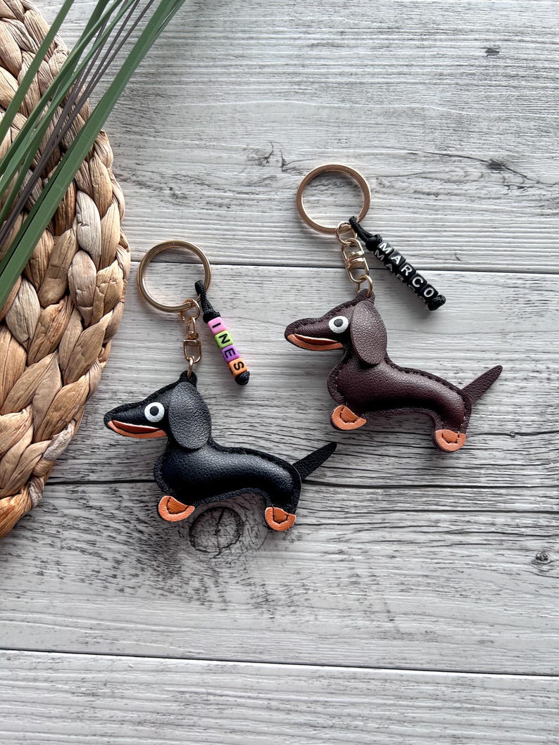 May include: Two dachshund keychains, one black and one brown, with orange accents on their paws and mouths. Each keychain has a gold ring and a beaded name tag. One tag says "INES" and the other says "MARCO".