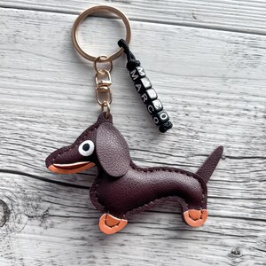 May include: A burgundy leather dachshund keychain with orange accents on its paws and snout. The keychain has a gold-colored ring and a black beaded charm that spells out the name "MARCO".