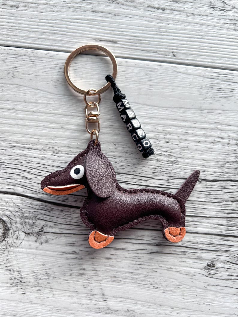 May include: A burgundy leather dachshund keychain with orange accents on its paws and snout. The keychain has a gold-colored ring and a black beaded charm that spells out the name "MARCO".
