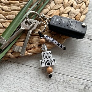 May include: A white keychain with the words "Dog Mom" and a paw print. The keychain has wooden beads and a silver charm. It is attached to a key ring with keys and a car key.