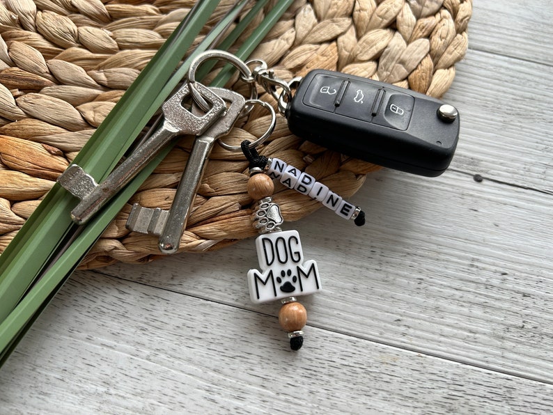 May include: A white keychain with the words "Dog Mom" and a paw print. The keychain has wooden beads and a silver charm. It is attached to a key ring with keys and a car key.