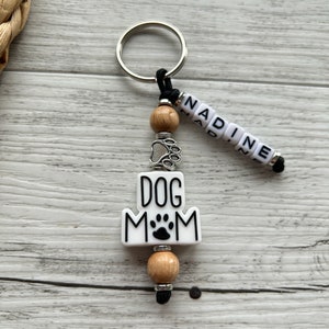 May include: White ceramic keychain with the text "DOG MOM" and a paw print. The keychain has a silver charm with a paw print, wooden beads, and a black cord with the name "NADINE" spelled out in white beads.