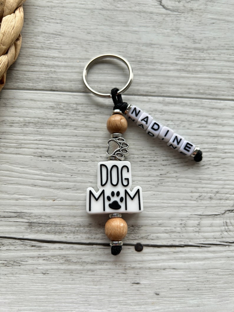 May include: White ceramic keychain with the text "DOG MOM" and a paw print. The keychain has a silver charm with a paw print, wooden beads, and a black cord with the name "NADINE" spelled out in white beads.