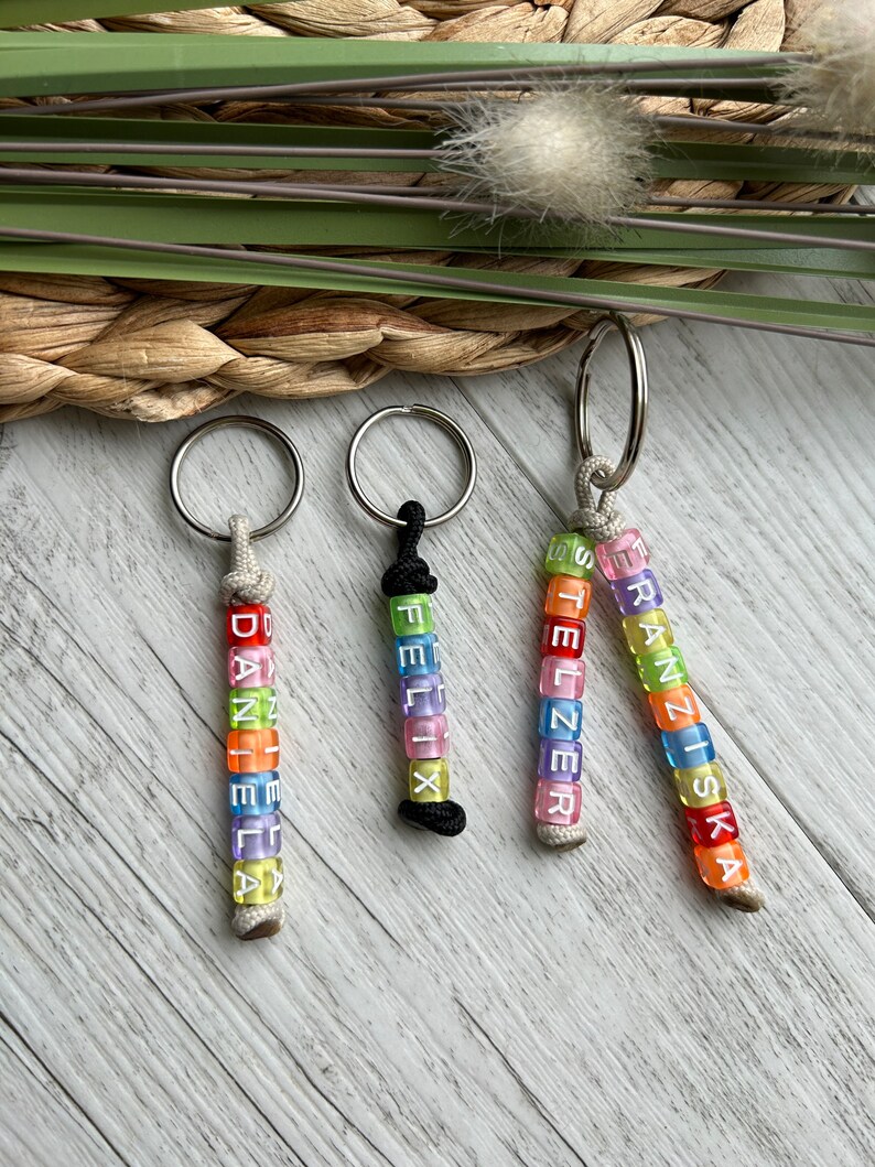 May include: Three keychains with names spelled out in colorful letter beads. The names are "DANIELA", "FELIX", and "FRANZISKA". Each keychain has a silver ring and a cord or string to hold the beads.