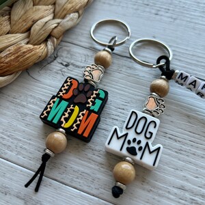 May include: Two keychains with wooden beads and metal rings. One keychain has a black and orange tag that says "Dog Mom" with a paw print. The other keychain has a white tag that says "Dog Mom" with a paw print.
