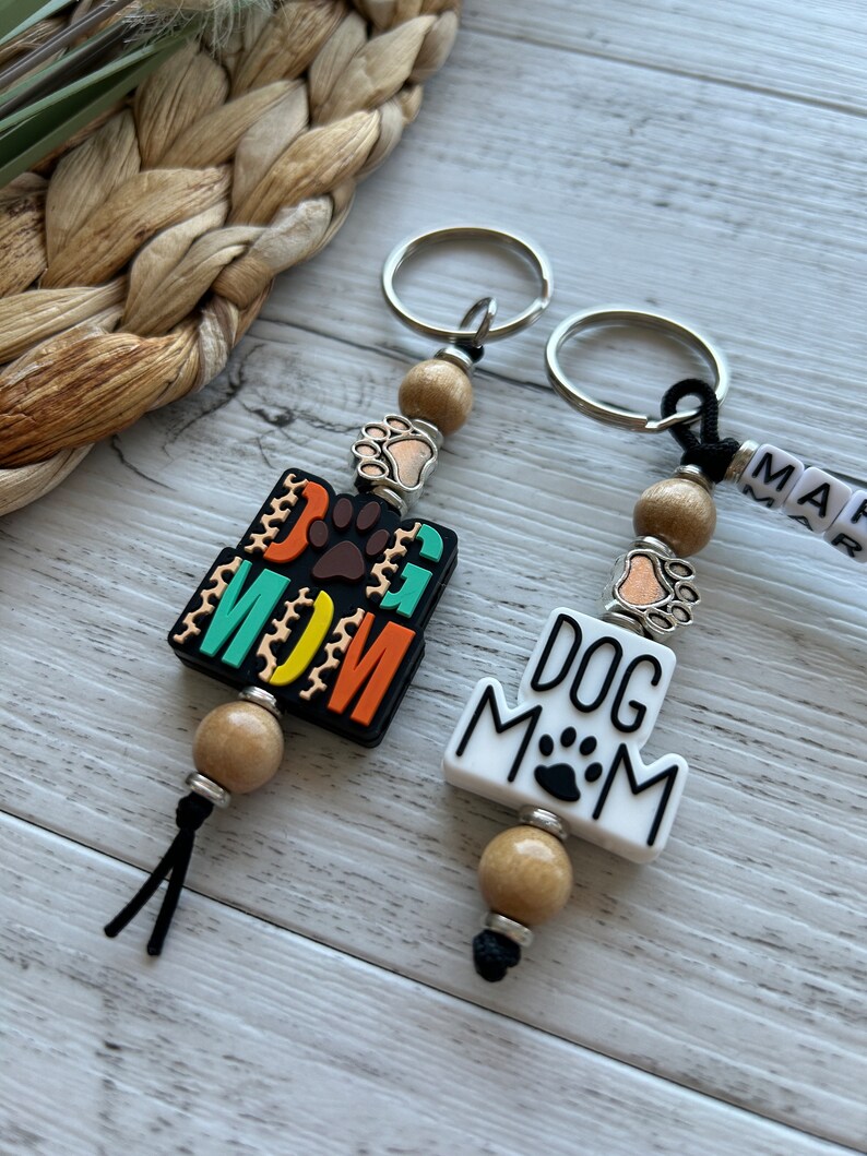 May include: Two keychains with wooden beads and metal rings. One keychain has a black and orange tag that says "Dog Mom" with a paw print. The other keychain has a white tag that says "Dog Mom" with a paw print.