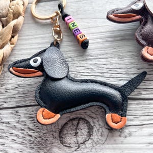 May include: A black leather dachshund keychain with orange accents on its paws and mouth. The keychain has a gold ring and a beaded charm spelling "INES". Another dachshund keychain is visible in the background.
