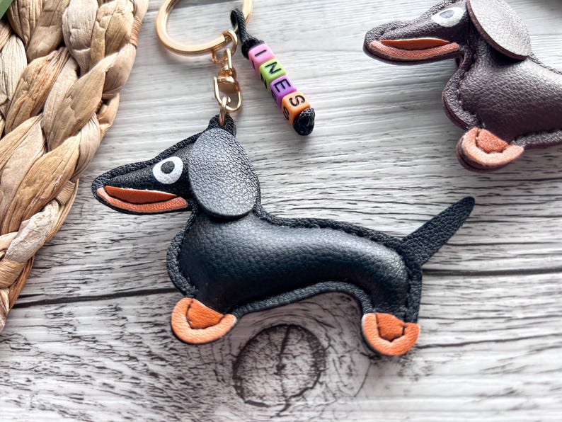 May include: A black leather dachshund keychain with orange accents on its paws and mouth. The keychain has a gold ring and a beaded charm spelling "INES". Another dachshund keychain is visible in the background.
