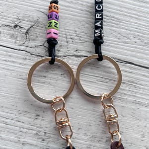 May include: Two keychains with gold-colored rings and clasps. One keychain has colorful letter beads spelling "SEZEN", the other has black letter beads spelling "MARCO". Both keychains have black cords.
