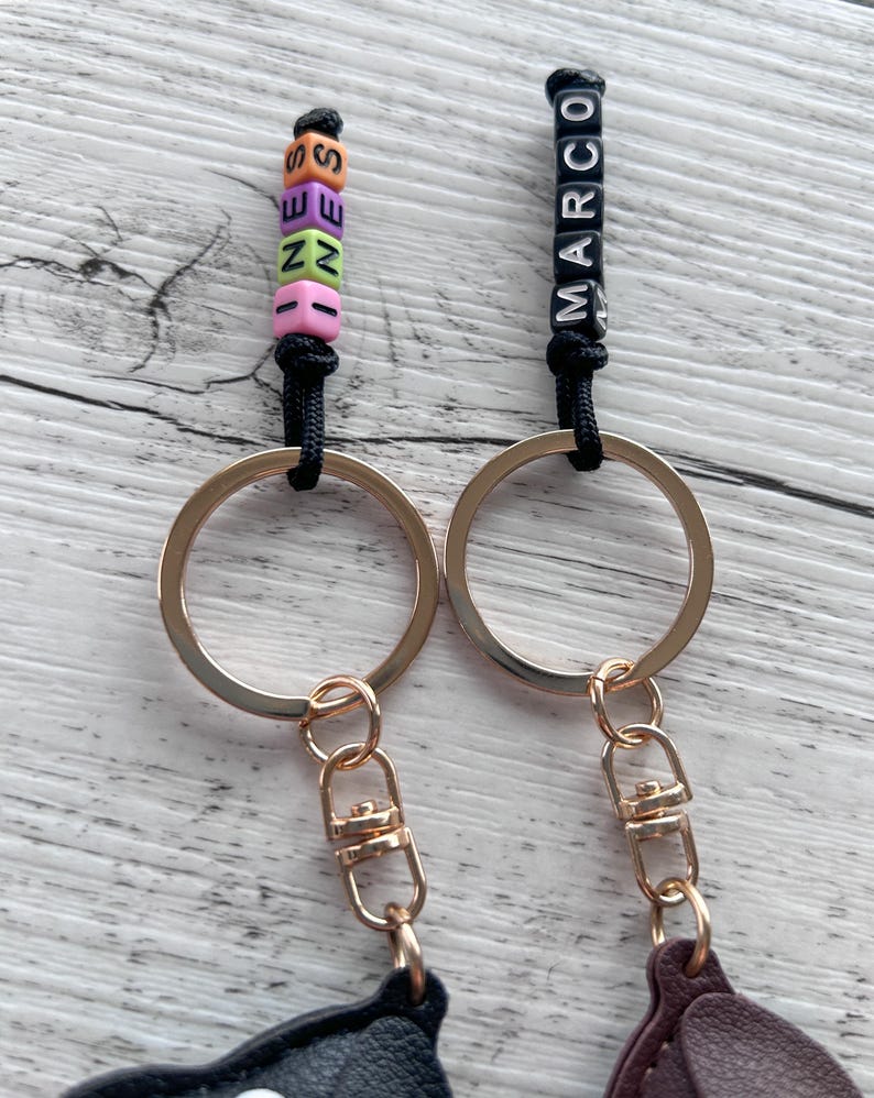 May include: Two keychains with gold-colored rings and clasps. One keychain has colorful letter beads spelling "SEZEN", the other has black letter beads spelling "MARCO". Both keychains have black cords.