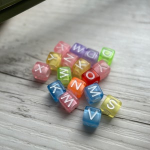 May include: A collection of colorful, translucent alphabet beads. Each cube-shaped bead displays a different letter in white, with a variety of colors including pink, blue, yellow, and green. The beads are scattered on a light-colored wooden surface.