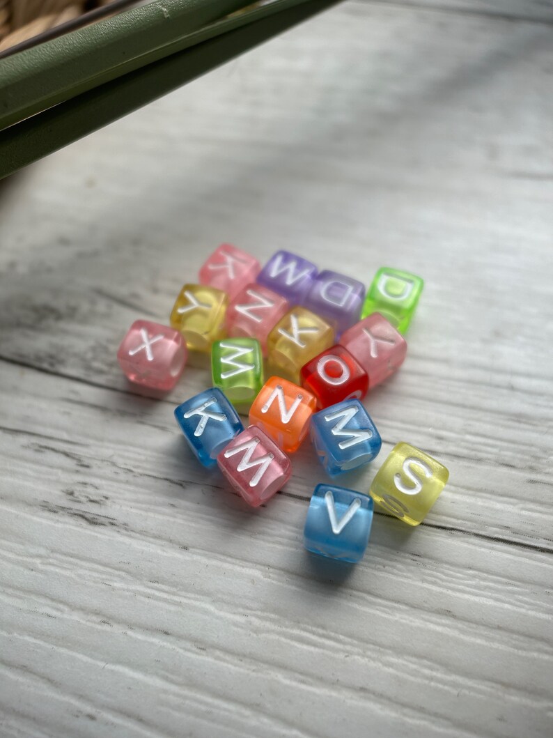 May include: A collection of colorful, translucent alphabet beads. Each cube-shaped bead displays a different letter in white, with a variety of colors including pink, blue, yellow, and green. The beads are scattered on a light-colored wooden surface.