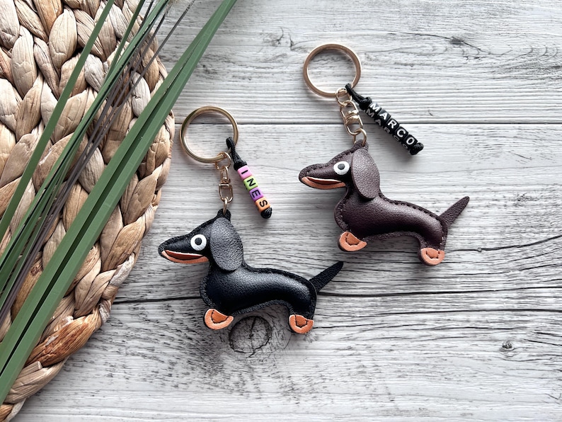 May include: Two dachshund keychains. One is black, the other is brown. Both have gold-colored key rings and orange-tan paws. One keychain has the name "INES" and the other has "MARCO" spelled out in colorful beads.
