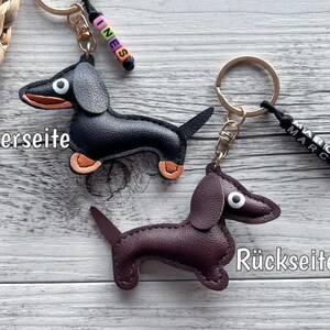 May include: Two dachshund keychains. One is black with orange paws, the other is brown. Each has a gold keyring and a small beaded name tag. The black keychain has the name "INES" and the brown keychain has the name "MARCO".