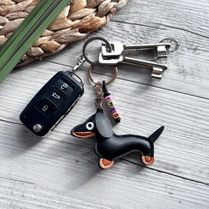 May include: A black leather dachshund keychain with tan accents, attached to a keyring with keys and a car remote. The keychain has a small beaded charm that spells out the name "INES". The items are on a light wood surface.