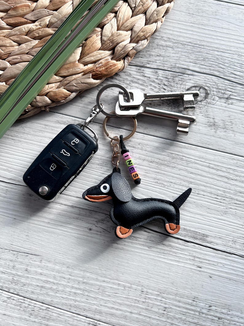 May include: A black leather dachshund keychain with tan accents, attached to a keyring with keys and a car remote. The keychain has a small beaded charm that spells out the name "INES". The items are on a light wood surface.