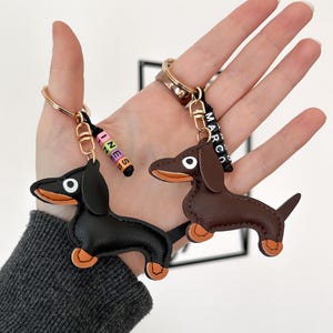 May include: Two dachshund keychains. One is black, the other is brown, both with orange paws and ears. Each keychain has a gold ring and a black strap with a name spelled out in colorful beads. One says "INES" and the other says "MARCO".