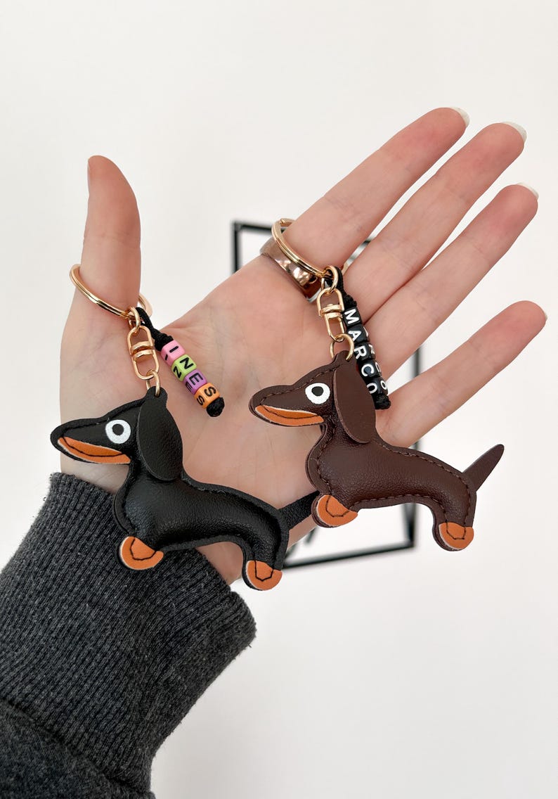 May include: Two dachshund keychains. One is black, the other is brown, both with orange paws and ears. Each keychain has a gold ring and a black strap with a name spelled out in colorful beads. One says "INES" and the other says "MARCO".