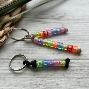 May include: Three keychains with rainbow-colored letter beads spelling out names. The top two keychains have names spelled out in clear, square beads with a silver keyring and a beige cord. The bottom keychain has the name "FELIX" in clear, square beads with a black cord.