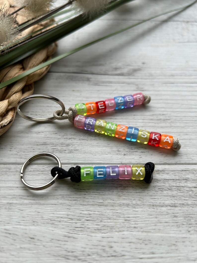 May include: Three keychains with rainbow-colored letter beads spelling out names. The top two keychains have names spelled out in clear, square beads with a silver keyring and a beige cord. The bottom keychain has the name "FELIX" in clear, square beads with a black cord.