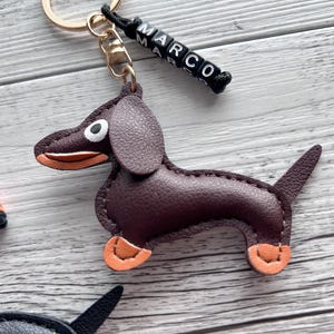 May include: A brown leather dachshund keychain with orange accents on its paws and mouth. The keychain has a gold-colored ring and a black cord with the name "MARCO" spelled out in white and black letter beads. The background is a light wood grain.