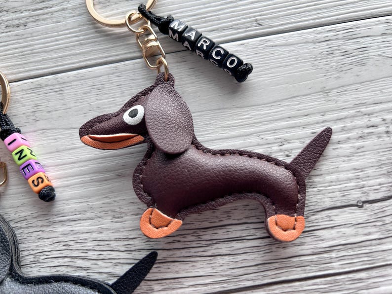May include: A brown leather dachshund keychain with orange accents on its paws and mouth. The keychain has a gold-colored ring and a black cord with the name "MARCO" spelled out in white and black letter beads. The background is a light wood grain.