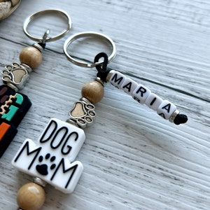 May include: Two keychains with brown wooden beads, silver paw print charms, and black cords. One keychain has a white charm with the text "DOG MOM" and a black paw print. The other keychain has white letter beads spelling "MARIA".