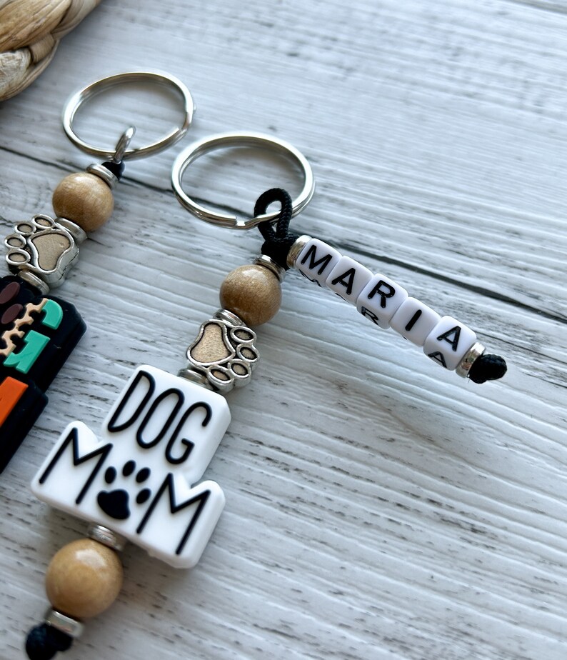 May include: Two keychains with brown wooden beads, silver paw print charms, and black cords. One keychain has a white charm with the text "DOG MOM" and a black paw print. The other keychain has white letter beads spelling "MARIA".