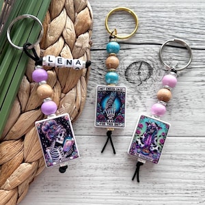 Könnte beinhalten: Drei bunte Schlüsselanhänger mit Holzperlen und Anhängern. Die Anhänger sind quadratisch und zeigen einen Schädel mit einer Krone, eine Hand mit einem Schädel und eine Kuh mit einem Schädel. Die Schlüsselanhänger tragen außerdem den Text "The Reader", "Not Enough Sage For This Shit" und "The Homebody".