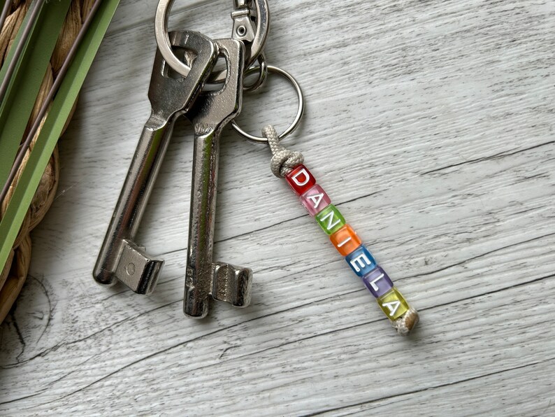 May include: A keychain with the name "DANIELA" spelled out in colorful square beads. The beads are in red, orange, green, blue, purple, and yellow. The keychain is attached to a silver keyring with two keys.