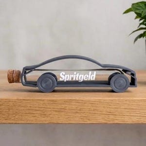 Könnte beinhalten: Eine graue Spardose in Autoform mit einem durchsichtigen Glasröhrenkörper, auf dem das Wort Spritgeld in weißen Buchstaben steht. Der Fahrzeugrahmen besteht aus grauem Material mit runden Rädern und einem Korken am hinteren Ende des Glasrohrs.