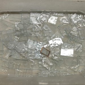 May include: A collection of clear glass pieces in various rectangular and square shapes, featuring diverse embossed textures such as grids, ripples, and floral patterns, alongside a single small rectangular frame with a metallic copper-colored edge.