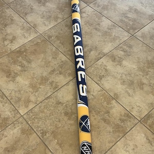 May include: A cylindrical, fabric-covered item with the word "SABRES" in white on a navy blue background. It has yellow and navy blue accents, with the NHL logo and a team logo. Likely a hockey-related product.