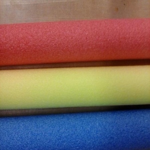 May include: Three colorful foam pool noodles in red, yellow, and blue. The cylindrical tubes are made of a textured foam material. The image shows a close-up of the pool noodles, with the colors arranged vertically.