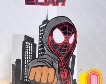 May include: A cake topper with a superhero theme. It features a red and black mask, a grey hooded sweatshirt, and a clenched fist. The name "ELIAH" is displayed in red letters. A cityscape silhouette is visible in the background. The words "partyplay" are also visible.