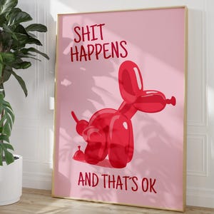 May include: A framed pink art print featuring a red balloon dog illustration. The text "SHIT HAPPENS AND THAT'S OK" is displayed in red. The artwork is displayed on a light wood floor.
