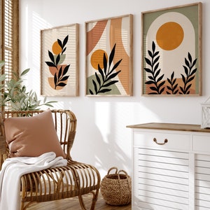 Puede incluir: Tres impresiones de arte abstracto enmarcadas con un dise&ntilde;o minimalista que presenta hojas estilizadas y formas geom&eacute;tricas en tonos marrones, naranjas y verdes. Las impresiones est&aacute;n colgadas en una pared blanca sobre un armario blanco con una tapa de madera.