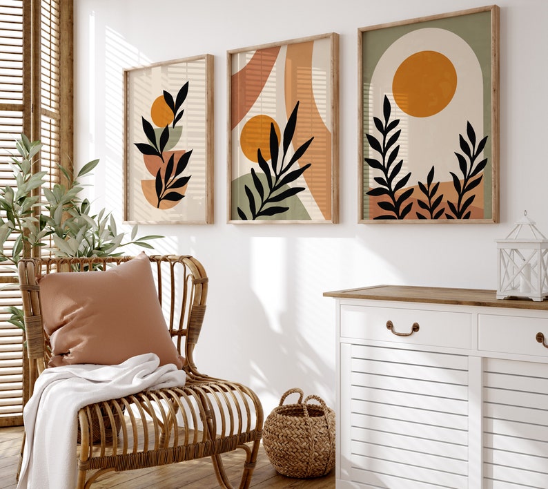 Puede incluir: Tres impresiones de arte abstracto enmarcadas con un dise&ntilde;o minimalista que presenta hojas estilizadas y formas geom&eacute;tricas en tonos marrones, naranjas y verdes. Las impresiones est&aacute;n colgadas en una pared blanca sobre un armario blanco con una tapa de madera.