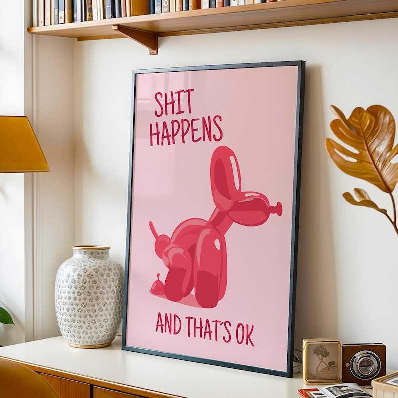 May include: A framed pink poster with the text "SHIT HAPPENS AND THAT'S OK" and a red balloon dog illustration. The poster is displayed on a white surface with a decorative vase and other items.